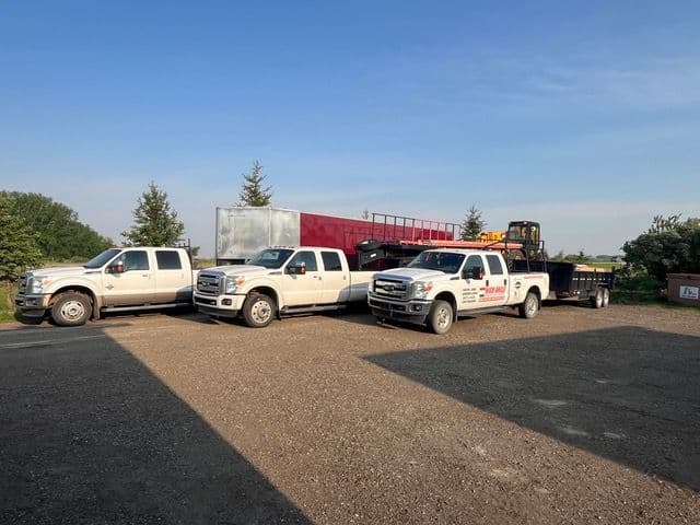 Four white trucks lined up with a trailer in an outdoor setting under blue skies.
