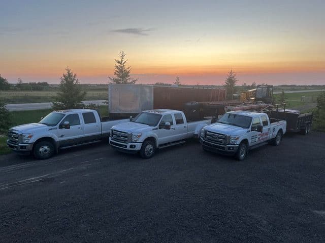 Three white pickup trucks parked near a sunset, showcasing a rural landscape and cargo trailers.