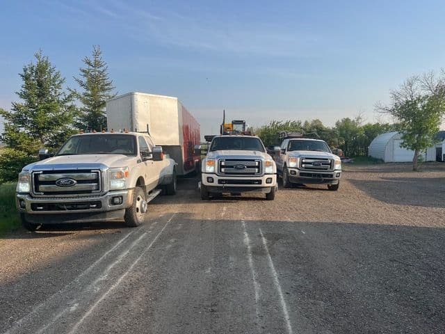 Three white Ford trucks parked on a gravel driveway with a trailer in the background.