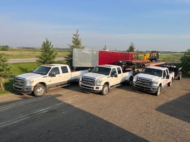 Three white pickup trucks parked in a gravel lot with a flatbed trailer and machinery in the background.